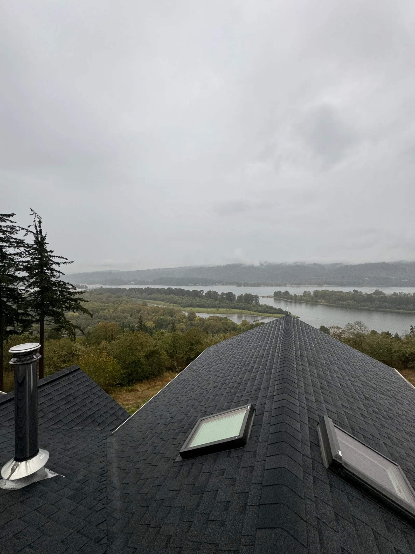 Charcoal gray dimensional shingle roof with skylight and metal chimney flashing overlooking water and forest landscape
