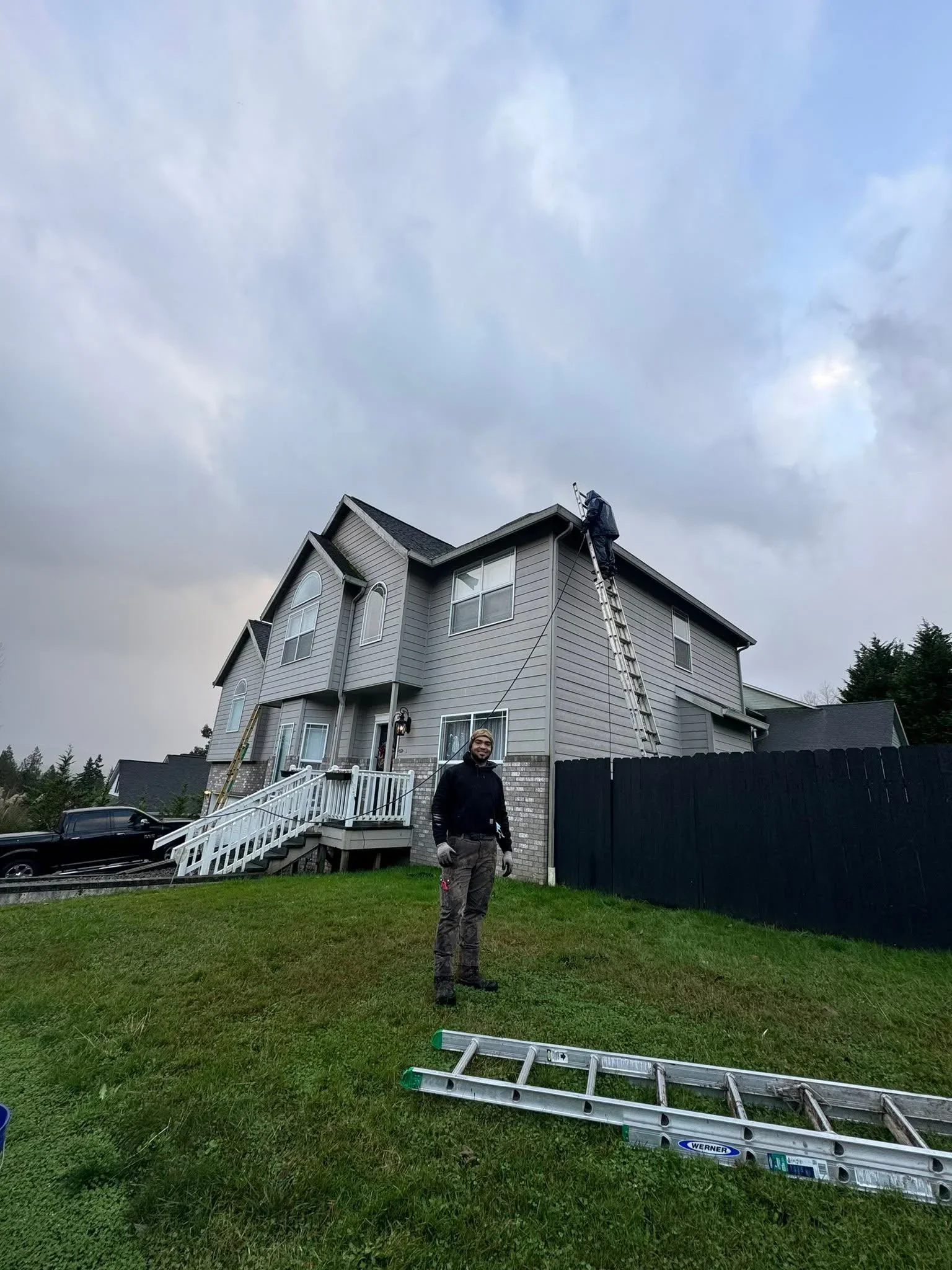 Roofer on extension ladder inspecting two-story home with gray siding and architectural shingles
